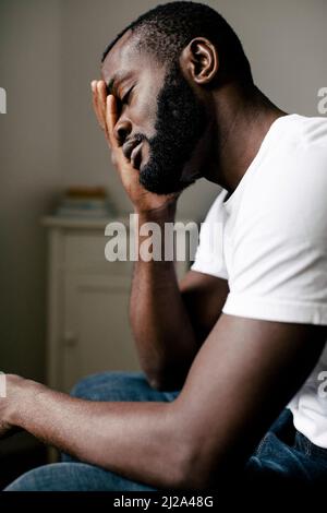 Side view of bearded lonely man covering closed eyes with hand sitting at home Stock Photo