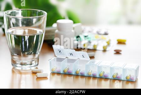 Daily Pill box organizer of vitamins and medicines with glass of water on the table. Stock Photo