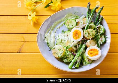 High angle of bowl with tasty vegetable spring salad made of asparagus and boiled eggs with potatoes served on wooden table with flowers Stock Photo