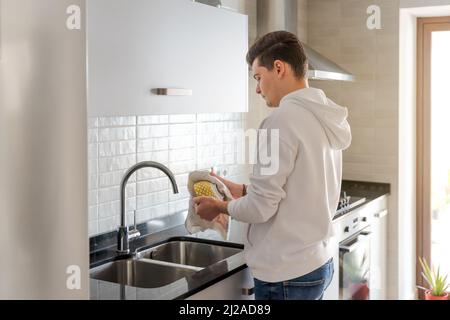 Young Caucasian man drying dishes with a cloth in a modern kitchen. White hoodie, casual lifestyle. Interior with natural light and white cabinets. Stock Photo