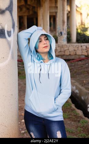 Smiling Teenager wearing light blue oversize hoodie and jeans stays under bridge . Blue haired teen girl stays outdoors against bridge pillars. Clothi Stock Photo