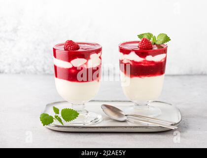 Two glasses with cold layered dessert. Raspberry sauce and buttermilk cream decorated with fresh berries and lemon balm leaves garnish. Stock Photo