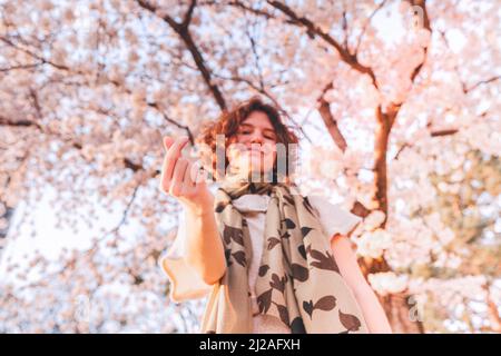 Lovely happy teen girl with curly brown hair, showing fingers Korean sign symbol heart, on blurred background of cherry blossoms. soft selective focus Stock Photo
