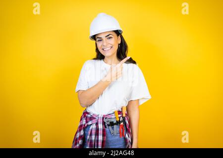 Young caucasian woman wearing hardhat and builder clothes over isolated yellow background smiling and pointing with hand and finger to the side Stock Photo