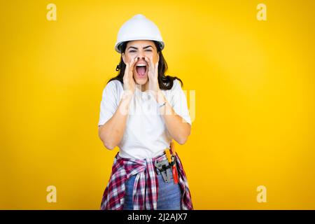 Young caucasian woman wearing hardhat and builder clothes over isolated yellow background shouting and screaming loud to side with hands on mouth Stock Photo