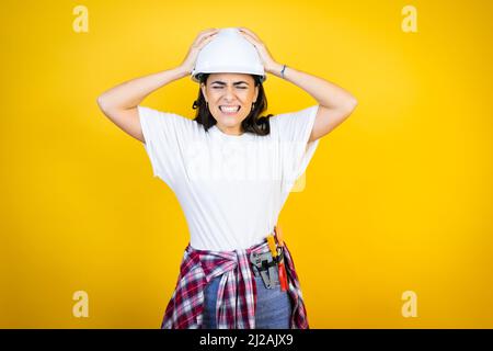 Young caucasian woman wearing hardhat and builder clothes over isolated yellow background suffering from headache desperate and stressed because pain Stock Photo