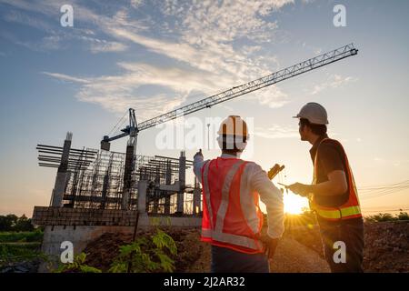 Asian two construction engineers supervising progress of construction project and crane background. Stock Photo