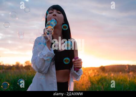 pretty young woman making soap bubbles in summer green field on sunset. happy peaceful time. millennial generation. Stock Photo