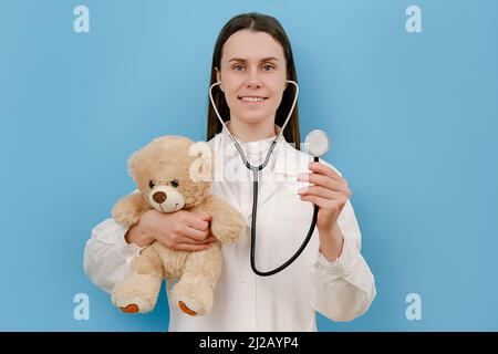 Portrait of cute smiling young woman doctor holding stethoscope and small fluffy bear, looking at camera, isolated over blue color background wall in Stock Photo