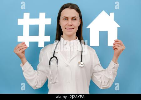 Happy young woman physician practitioner in white uniform with stethoscope holds hashtag sign and house model, posing on blue color background. Corona Stock Photo