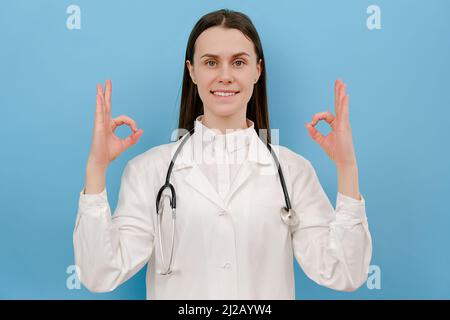 Cute smiling young female doctor in white coat with stethoscope showing ok sign and thumbs up, positive looking at camera, isolated over blue color wa Stock Photo