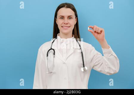 Woman cardiologist in white coat showing to camera cardiac medication pill, isolated on blue background. Vitamin complex sale discount, effective trea Stock Photo