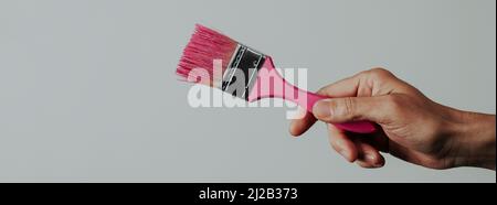a young man grabs a paintbrush with some pink paint, on a pale gray background, in a panoramic format to use as web banner or header Stock Photo