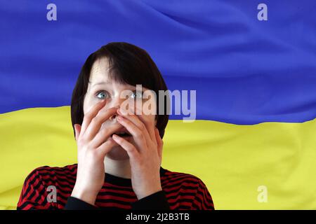 frightened teenage girl closes her mouth with her hands against the background of the Ukrainian flag. Stock Photo