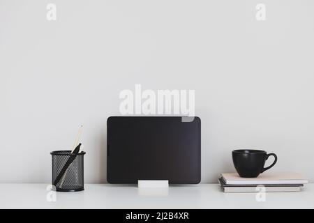 Digital tablet on holder, mug of tea, office stationery on white table. Home office, modern workspace. Stock Photo