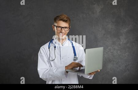 Portrait of happy male healthcare worker standing with laptop in hands on gray background. Stock Photo