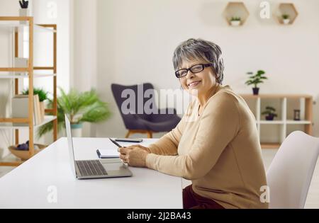 Joyful smiling confident senior woman using laptop computer sitting at workplace desk. Stock Photo