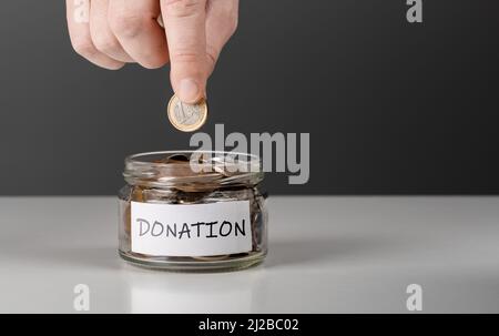 Man putting money into donation jar on table against grey background, closeup. Space for text. Hand putting Coins in glass jar for giving and donation Stock Photo