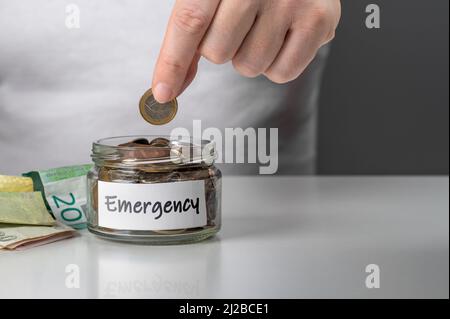 hand puts one euro coin in jar with the word emergency. hand putting coin in the glass jar for saving and emergency, Saving for Emergency concept. Stock Photo