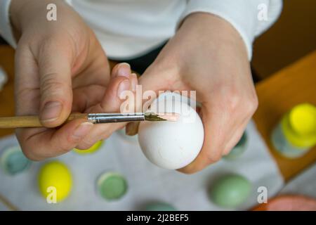 Holding paint brush in woman's hands, a clean white chicken egg view from above. A woman prepares to start painting egg with a beige paint. DIY eggs E Stock Photo