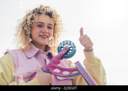 Pretty young blond curly haired woman shows thumb up to the camera and holds a scooter, smiling medium closeup copy space . High quality photo Stock Photo