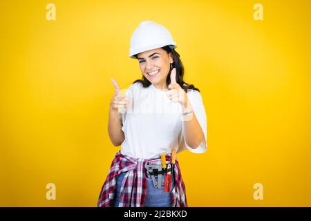 Young caucasian woman wearing hardhat and builder clothes over isolated yellow background pointing to you and the camera with fingers, smiling positiv Stock Photo