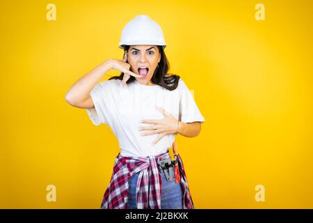 Young caucasian woman wearing hardhat and builder clothes over isolated yellow background confused doing phone gesture with hand and fingers like talk Stock Photo