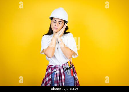 Young caucasian woman wearing hardhat and builder clothes over isolated yellow background sleeping tired dreaming and posing with hands together while Stock Photo