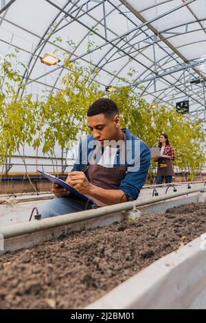 African american farmer writing on clipboard near ground in greenhouse Stock Photo