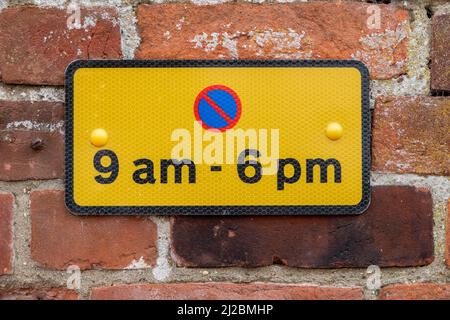 Small rectangular parking restriction sign black on yellow with red and blue symbol for the hours of 9am to 6pm mounted on a red brick wall Stock Photo