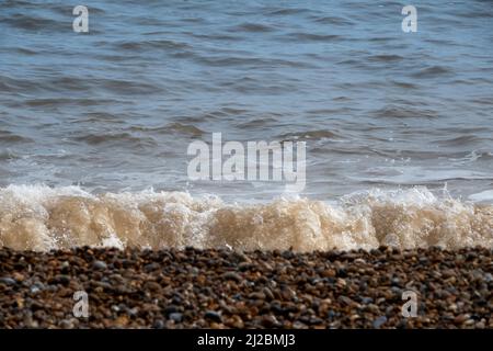 Small waves breaking on the shoreline of a pebble beach with bubbly white foam and soft focus beach in the foreground Stock Photo