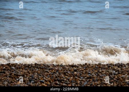 Small waves breaking on the shoreline of a pebble beach with bubbly white foam and soft focus beach in the foreground Stock Photo