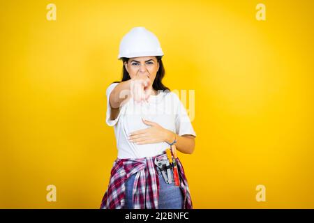 Young caucasian woman wearing hardhat and builder clothes over isolated yellow background laughing at you, pointing finger to the camera with hand ove Stock Photo