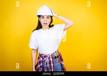 Young caucasian woman wearing hardhat and builder clothes over isolated yellow background putting one hand on her head smiling like she had forgotten Stock Photo