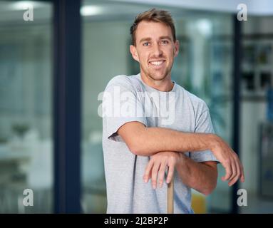 Need something cleaned Im your man. Portrait of a young man leaning on a mop while cleaning the office floor. Stock Photo