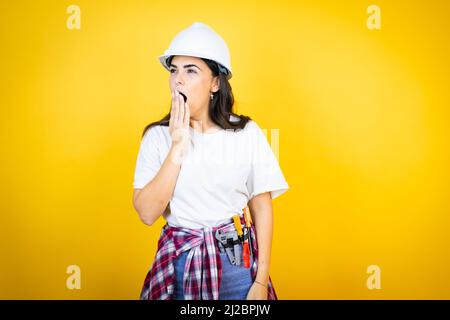 Young caucasian woman wearing hardhat and builder clothes over isolated yellow background bored yawning tired covering mouth with hand. Restless and s Stock Photo