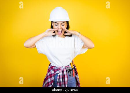 Young caucasian woman wearing hardhat and builder clothes over isolated yellow background shouting and screaming loud down with hands on mouth Stock Photo