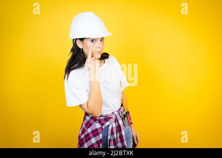 Young caucasian woman wearing hardhat and builder clothes over isolated yellow background Pointing to the eye watching you gesture, suspicious express Stock Photo