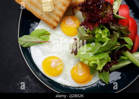 Breakfast. Fried eggs with toast with butter and vegetables, cucumbers and tomatoes. On a dark plate, against a dark background Stock Photo
