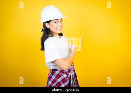 Young caucasian woman wearing hardhat and builder clothes over isolated yellow background feeling happy, positive and successful, motivated when facin Stock Photo