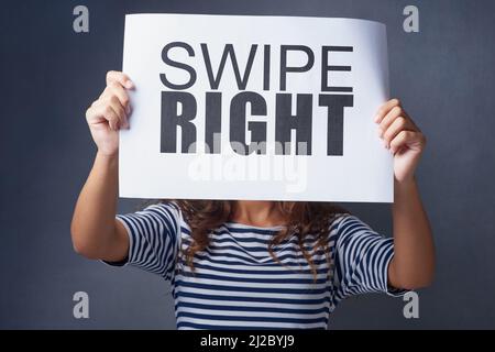 Its a match. Studio shot of a young woman holding a sign with swipe right printed on it against a gray background. Stock Photo
