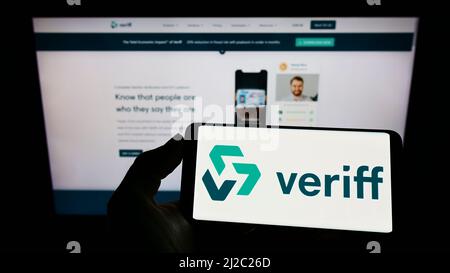 Person holding cellphone with logo of Estonian identity verification company Veriff on screen in front of webpage. Focus on phone display. Stock Photo