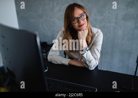 A beautiful elderly woman with black glasses sits at a desk, looks at the camera, and holds her left hand on her chin. She is in her office. Stock Photo