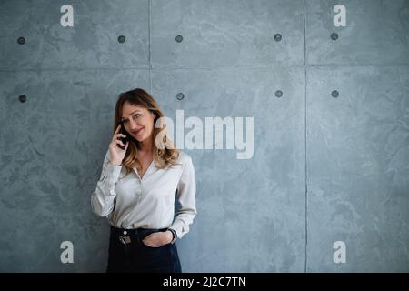 Very complacent,fashionable,beauteous,assertive woman lawyer in her 50s is making phone call in her modern, gray office.She's holding her hand in pocket Stock Photo