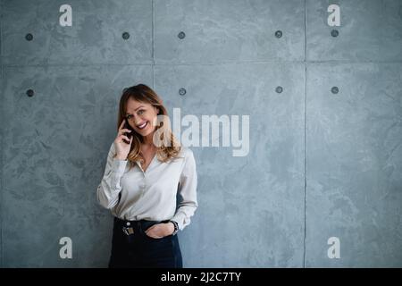 A Caucasian woman stands in front of gray background in the office with her left hand in her pocket. She looks at the camera and has a phone call. Stock Photo