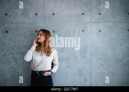 Adult woman in office makes important phone call.Left hand is in pocket.Gray,fashionable wall is just behing her.She stands and looks to the door. Stock Photo