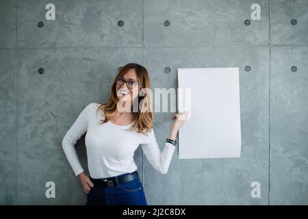 Caucasian middle-aged smiling woman with spectacles is pointing to white paper on the gray wall on her left side. She is presenting in denim and shirt Stock Photo