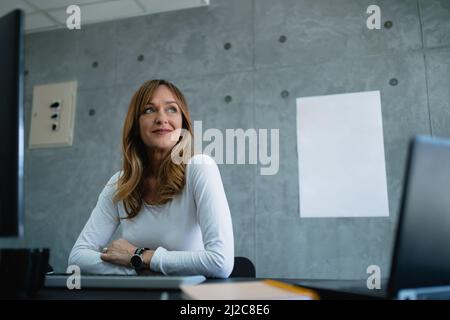 A beautiful green eye senior woman is sitting in front of white paper which is on gray background. There are desktop and laptop on her desk. Stock Photo