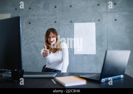 Caucasian female with the raised thumb is showing OK in office. A middle-aged, brown-haired woman trainer in a white shirt is sitting at her desk. Stock Photo