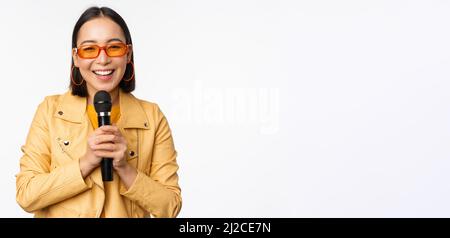 Stylish asian girl in sunglasses, singing songs with microphone, holding mic and dancing at karaoke, posing against white background Stock Photo